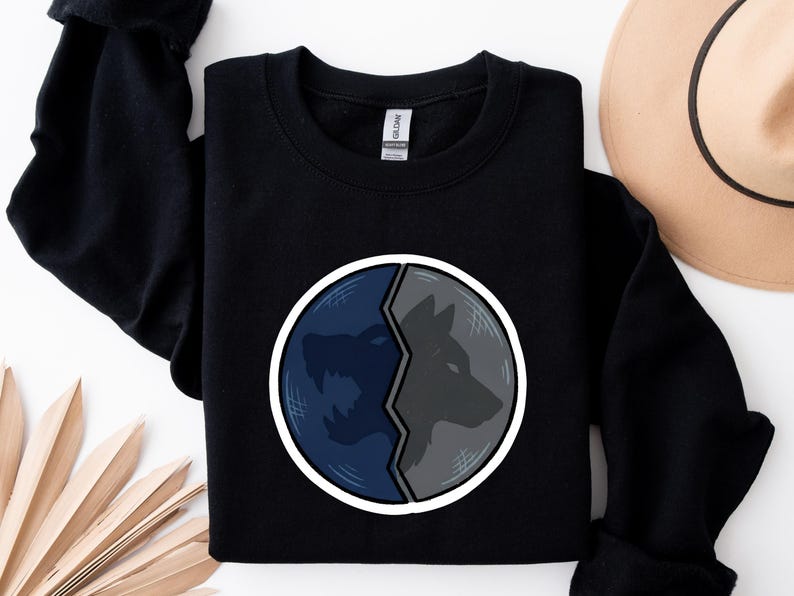 Puede incluir: Sudadera negra con un dise&ntilde;o circular dividido en dos. Un lado muestra un lobo azul aullando, el otro un perfil de lobo gris. El dise&ntilde;o est&aacute; delineado en blanco, creando un contraste llamativo.