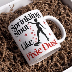 May include: White ceramic mug with the text "Sprinkling Smut Like Pixie Dust" in black and red. A silhouette of a woman is depicted, sprinkling stars. The mug is inside a white box with brown paper filler.