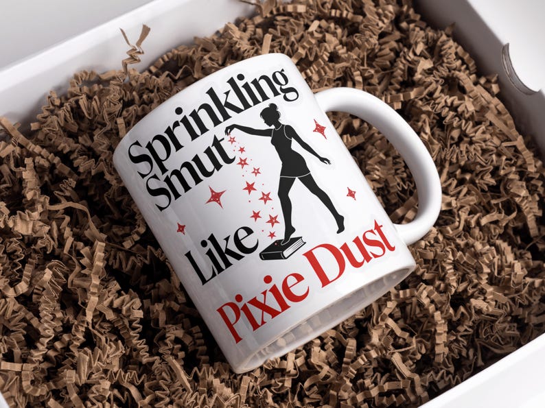 May include: White ceramic mug with the text "Sprinkling Smut Like Pixie Dust" in black and red. A silhouette of a woman is depicted, sprinkling stars. The mug is inside a white box with brown paper filler.