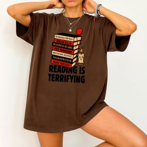 May include: A brown t-shirt with a graphic design featuring a stack of books with titles like "Dracula" and "Frankenstein," a candle, and the text "Reading is Terrifying."