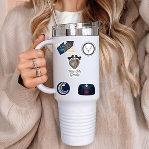 Puede incluir: Vaso t&eacute;rmico blanco con tapa plateada, adornado con varias pegatinas. Las pegatinas incluyen una luna creciente, un coraz&oacute;n con lazos y el texto "Bite Me Gently". El vaso es sostenido por una persona que lleva un su&eacute;ter beige.