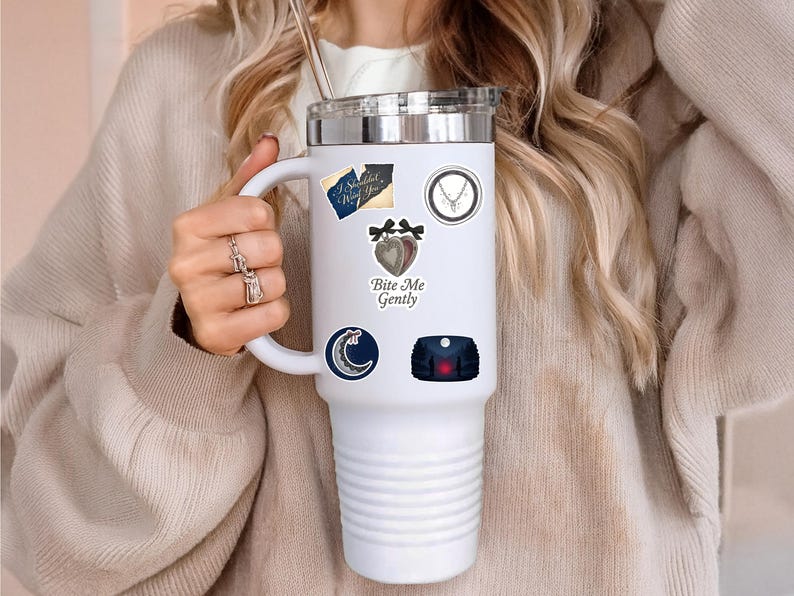 Puede incluir: Vaso t&eacute;rmico blanco con tapa plateada, adornado con varias pegatinas. Las pegatinas incluyen una luna creciente, un coraz&oacute;n con lazos y el texto "Bite Me Gently". El vaso es sostenido por una persona que lleva un su&eacute;ter beige.