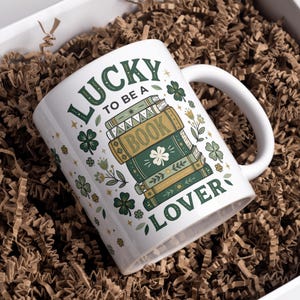 May include: White ceramic mug with the text "LUCKY TO BE A BOOK LOVER" in green lettering. The mug features a stack of books with a shamrock and floral accents. The mug is inside a white box with brown paper filler.