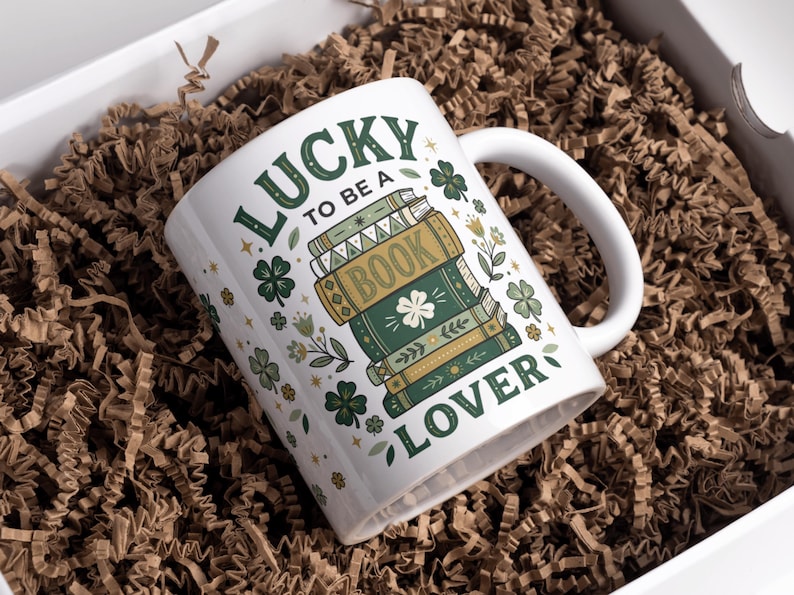 May include: White ceramic mug with the text "LUCKY TO BE A BOOK LOVER" in green lettering. The mug features a stack of books with a shamrock and floral accents. The mug is inside a white box with brown paper filler.