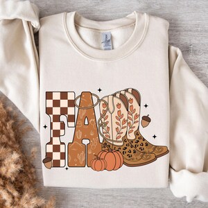 May include: A cream-colored crewneck sweatshirt featuring a graphic print that spells FALL. The F is a brown and white checkerboard, the A contains cactus illustrations, and the LL are depicted as two cowboy boots with leopard print accents.