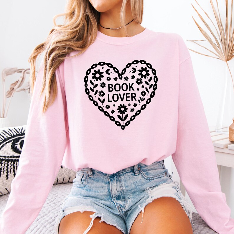 Puede incluir: Sudadera rosa claro con un dise&ntilde;o de coraz&oacute;n negro que dice "BOOK LOVER". El coraz&oacute;n est&aacute; decorado con flores y hojas. La sudadera tiene mangas largas.