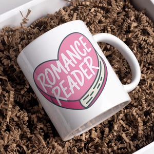 May include: White ceramic mug with a pink heart design that reads "ROMANCE READER" in white text. The mug is inside a white box filled with brown crinkle paper.