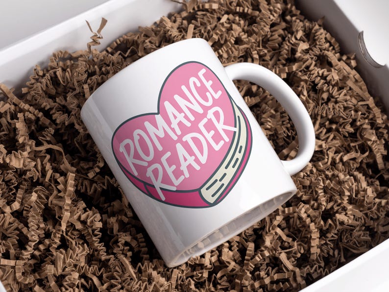 May include: White ceramic mug with a pink heart design that reads "ROMANCE READER" in white text. The mug is inside a white box filled with brown crinkle paper.