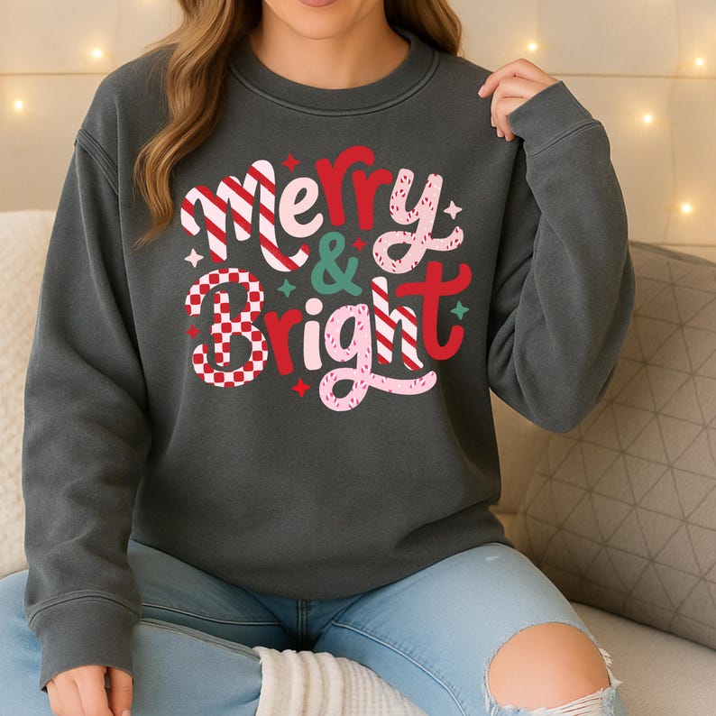 以下が含まれることがあります： 「Merry & Bright」の文字がデザインされた、お祝いの雰囲気のダークグレーのスウェットシャツ。赤、ピンク、白の色使いと星のアクセントが特徴で、ホリデーシーズンにぴったりです。クルーネックのスタイルです。