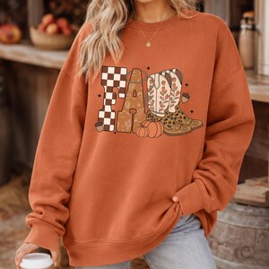 May include: A burnt orange crewneck sweatshirt featuring a graphic print on the chest. The design displays the word FALL in large letters, with the F in a brown and white checkerboard pattern, and the A, L, and L decorated with cowboy boots, a pumpkin, and acorns.
