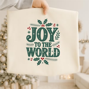 May include: Cream-colored tote bag with the text "JOY TO THE WORLD" in dark green, accented with holly and berries. The bag is held up by a person. Festive holiday-themed design.