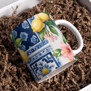 May include: A white ceramic mug with a vibrant design featuring lemons, pink flowers, and blue floral patterns. The mug is set inside a white box filled with brown crinkle paper, suggesting a gift presentation.