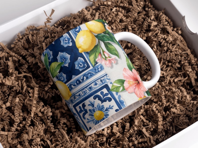 May include: A white ceramic mug with a vibrant design featuring lemons, pink flowers, and blue floral patterns. The mug is set inside a white box filled with brown crinkle paper, suggesting a gift presentation.