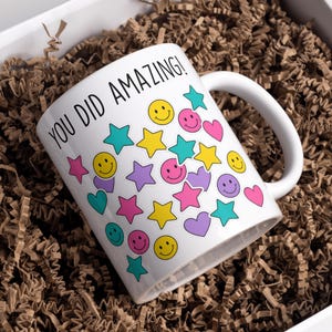 May include: White ceramic mug with the text "YOU DID AMAZING!" and colorful smiley faces, stars, and hearts. The mug is inside a white box filled with brown crinkle paper.