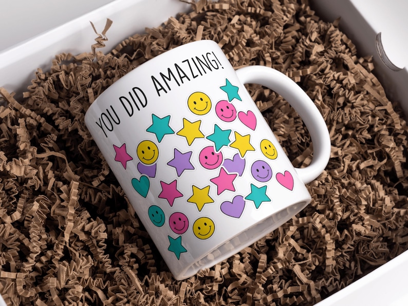 May include: White ceramic mug with the text "YOU DID AMAZING!" and colorful smiley faces, stars, and hearts. The mug is inside a white box filled with brown crinkle paper.