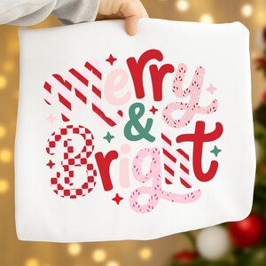 以下が含まれることがあります： 赤、ピンク、緑で「Merry & Bright」と書かれた白い布。テキストは、星とひし形模様の、お菓子のようなお祭りデザインです。全体的なテーマはホリデーデコレーションです。