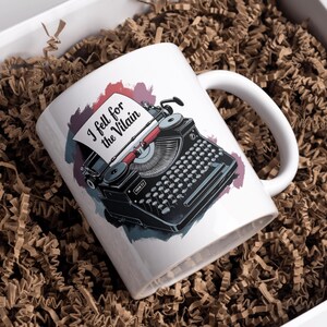 Puede incluir: Taza de cer&aacute;mica blanca con una ilustraci&oacute;n de m&aacute;quina de escribir vintage negra y el texto "I fell for the Villain". La taza est&aacute; en una caja blanca con papel arrugado marr&oacute;n.