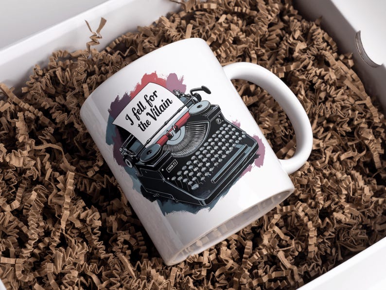 Puede incluir: Taza de cer&aacute;mica blanca con una ilustraci&oacute;n de m&aacute;quina de escribir vintage negra y el texto "I fell for the Villain". La taza est&aacute; en una caja blanca con papel arrugado marr&oacute;n.