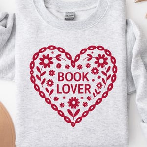 Puede incluir: Una sudadera gris claro con un dise&ntilde;o de coraz&oacute;n rojo. El coraz&oacute;n est&aacute; decorado con patrones florales y de hojas, y las palabras "BOOK LOVER" est&aacute;n impresas en el centro. La sudadera tiene cuello redondo y mangas largas.