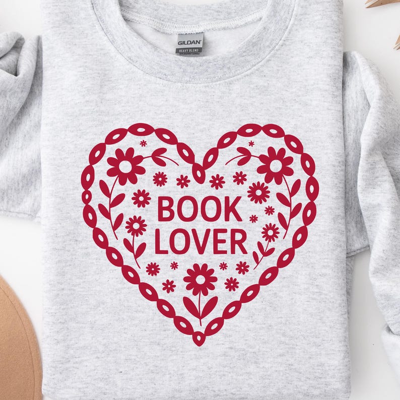 Puede incluir: Una sudadera gris claro con un dise&ntilde;o de coraz&oacute;n rojo. El coraz&oacute;n est&aacute; decorado con patrones florales y de hojas, y las palabras "BOOK LOVER" est&aacute;n impresas en el centro. La sudadera tiene cuello redondo y mangas largas.