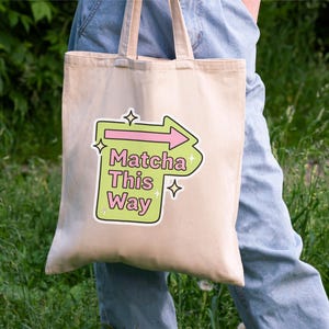 Puede incluir: Un bolso tote beige con el texto "Matcha This Way" en un gr&aacute;fico verde y rosa. El bolso tiene un asa de color claro y se sostiene sobre un fondo de follaje verde y vaqueros azules.