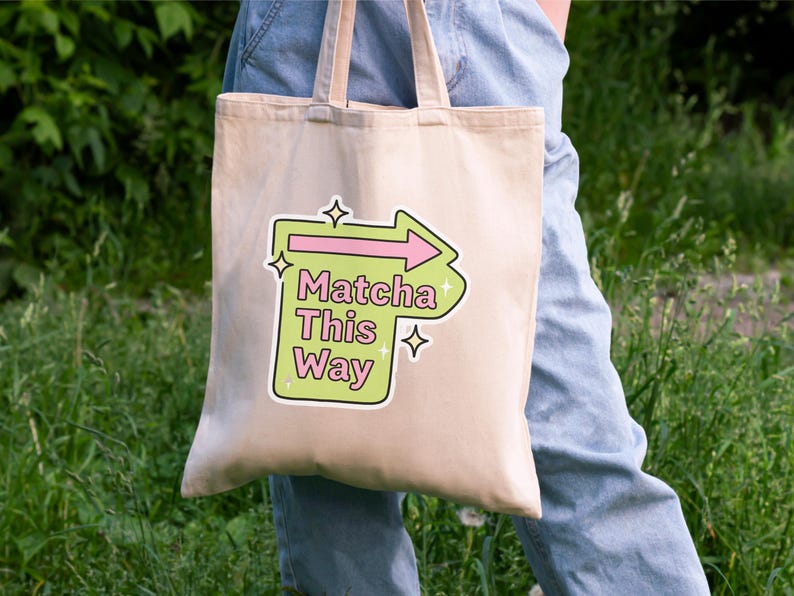 Puede incluir: Un bolso tote beige con el texto "Matcha This Way" en un gr&aacute;fico verde y rosa. El bolso tiene un asa de color claro y se sostiene sobre un fondo de follaje verde y vaqueros azules.