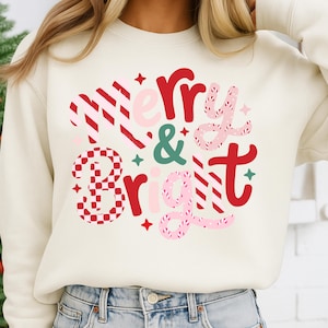 以下が含まれることがあります： クリーム色のスウェットシャツに「Merry & Bright」の文字が赤、ピンク、緑で描かれています。テキストはストライプ、チェック、星のアクセントが施されたお祝いのデザインで、ホリデーシーズンに最適です。