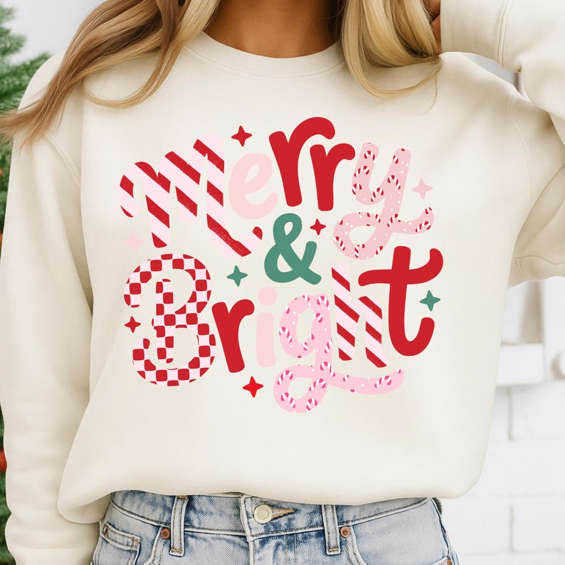 以下が含まれることがあります： クリーム色のスウェットシャツに「Merry & Bright」の文字が赤、ピンク、緑で描かれています。テキストはストライプ、チェック、星のアクセントが施されたお祝いのデザインで、ホリデーシーズンに最適です。