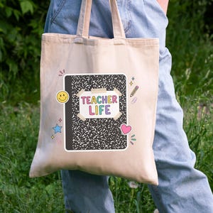 May include: A beige tote bag with a black and white composition notebook design. The notebook has the words "TEACHER LIFE" in colorful letters. The bag also features a yellow smiley face, a blue star, a pink heart, and a pencil.