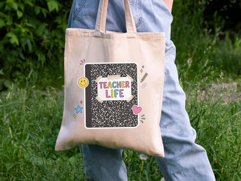 May include: A beige tote bag with a black and white composition notebook design. The notebook has the words "TEACHER LIFE" in colorful letters. The bag also features a yellow smiley face, a blue star, a pink heart, and a pencil.
