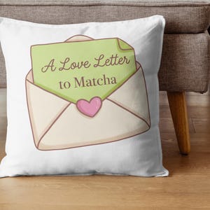 Puede incluir: Coj&iacute;n blanco con una ilustraci&oacute;n de dibujos animados de un sobre abierto con una carta verde que dice "A Love Letter to Matcha". Un coraz&oacute;n rosa est&aacute; debajo del texto. El coj&iacute;n est&aacute; sobre un suelo de madera.