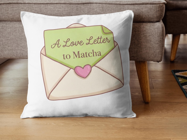 Puede incluir: Coj&iacute;n blanco con una ilustraci&oacute;n de dibujos animados de un sobre abierto con una carta verde que dice "A Love Letter to Matcha". Un coraz&oacute;n rosa est&aacute; debajo del texto. El coj&iacute;n est&aacute; sobre un suelo de madera.