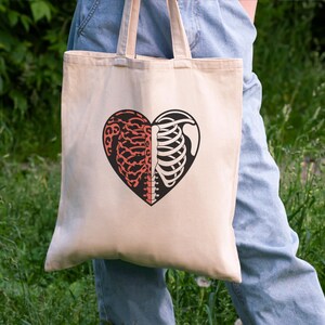 May include: Beige tote bag featuring a heart-shaped design split into two halves. One side depicts a brain in red, and the other side shows a rib cage in black. The bag has beige handles and is held against a background of green foliage.