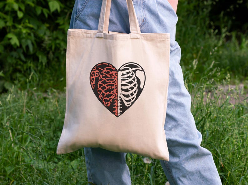 May include: Beige tote bag featuring a heart-shaped design split into two halves. One side depicts a brain in red, and the other side shows a rib cage in black. The bag has beige handles and is held against a background of green foliage.