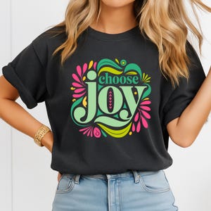 May include: Dark gray t-shirt with the words "choose Joy" in a colorful, floral design. The text is in a playful font, with green, pink, and yellow accents. The shirt has short sleeves and a relaxed fit.