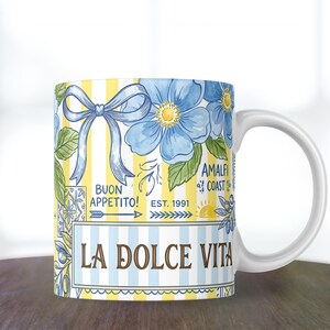May include: White ceramic mug with a colorful design. The mug features blue flowers, a blue bow, and the words "LA DOLCE VITA" and "BUON APPETITO!". The background has yellow and white stripes. The mug also has the words "AMALFI COAST EST. 1991".