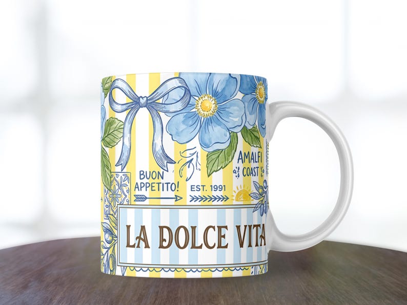 May include: White ceramic mug with a colorful design. The mug features blue flowers, a blue bow, and the words "LA DOLCE VITA" and "BUON APPETITO!". The background has yellow and white stripes. The mug also has the words "AMALFI COAST EST. 1991".