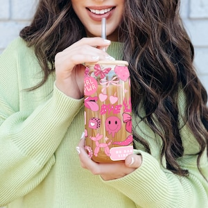 May include: A clear glass tumbler with a bamboo lid and a stainless steel straw. The tumbler is decorated with pink stickers, including hearts, lips, butterflies, and the words "Babe" and "No Bad Vibes". The person is wearing a light green sweater.