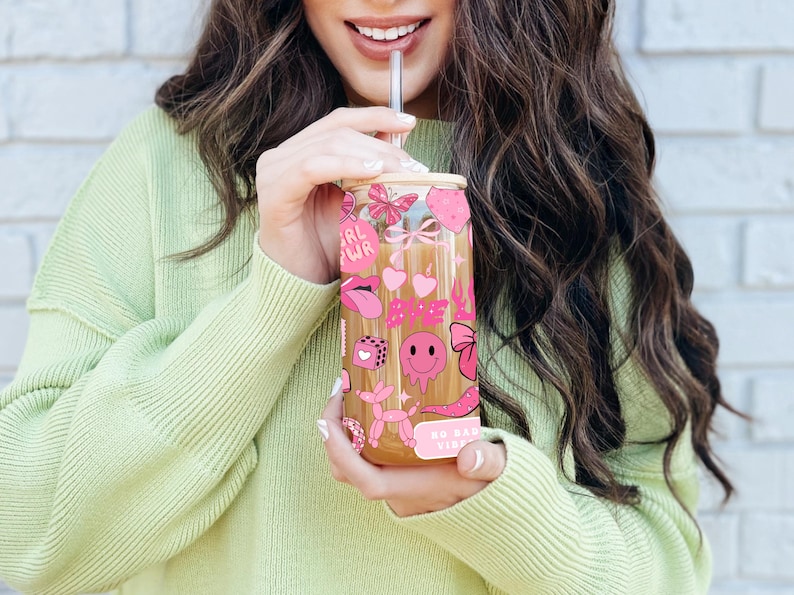 May include: A clear glass tumbler with a bamboo lid and a stainless steel straw. The tumbler is decorated with pink stickers, including hearts, lips, butterflies, and the words "Babe" and "No Bad Vibes". The person is wearing a light green sweater.