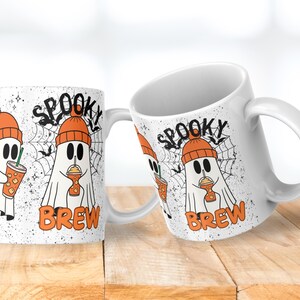 May include: White ceramic mug with a Halloween-themed design. The mug features a cartoon ghost wearing an orange beanie and holding a drink. The words "Spooky Brew" are printed on the mug, along with a spiderweb and bats.