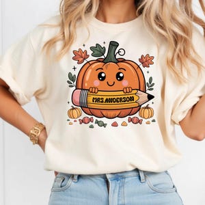May include: Cream-colored t-shirt featuring a cartoon pumpkin holding a pencil. The pumpkin has a smiling face and is surrounded by autumn leaves and small pumpkins. The pencil reads "MRS. ANDERSON".