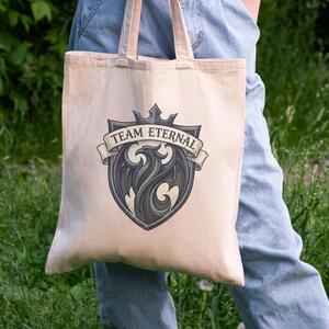 Puede incluir: Bolso tote beige con un emblema gris y negro que presenta un drag&oacute;n estilizado y el texto "TEAM ETERNAL" en una pancarta. La bolsa tiene asas beige a juego. El fondo es follaje verde.