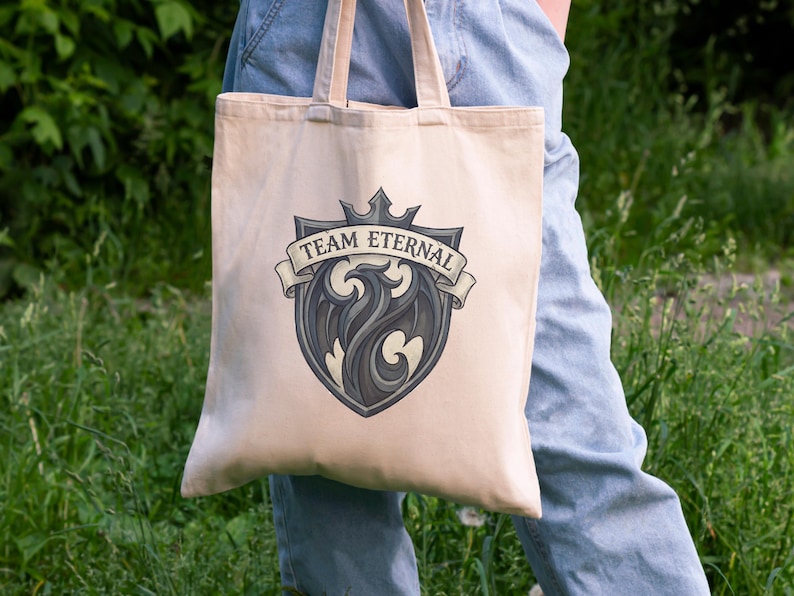 Puede incluir: Bolso tote beige con un emblema gris y negro que presenta un drag&oacute;n estilizado y el texto "TEAM ETERNAL" en una pancarta. La bolsa tiene asas beige a juego. El fondo es follaje verde.