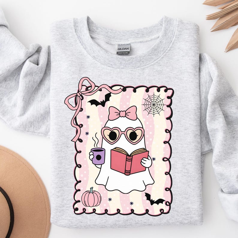 May include: Light gray sweatshirt featuring a cute ghost illustration. The ghost wears heart-shaped glasses and a bow, holding a book and a coffee cup. The design includes bats, a spiderweb, and a pumpkin, perfect for Halloween.