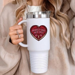 May include: White travel mug with a silver lid and handle. The mug features a red heart-shaped graphic with the text "Morally Gray Men Only" in gold lettering. The mug is held by a person wearing a beige sweater.