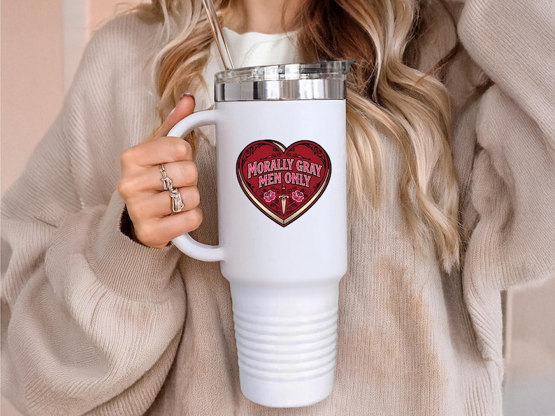 May include: White travel mug with a silver lid and handle. The mug features a red heart-shaped graphic with the text "Morally Gray Men Only" in gold lettering. The mug is held by a person wearing a beige sweater.