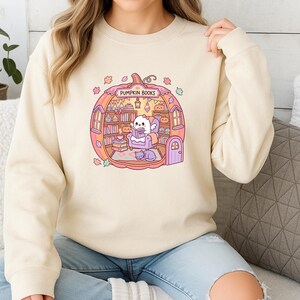 May include: Beige sweatshirt featuring a whimsical illustration of a ghost reading inside a pumpkin-shaped bookstore. The design includes bookshelves, a cat, and the text "PUMPKIN BOOKS" in a circular frame, with autumn leaves and pumpkins.