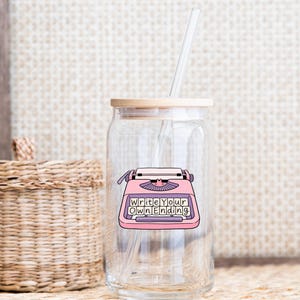 May include: A clear glass tumbler with a bamboo lid and a reusable straw. The tumbler features a pink typewriter graphic with the words "Write Your Own Ending". A woven basket and textured background are visible.