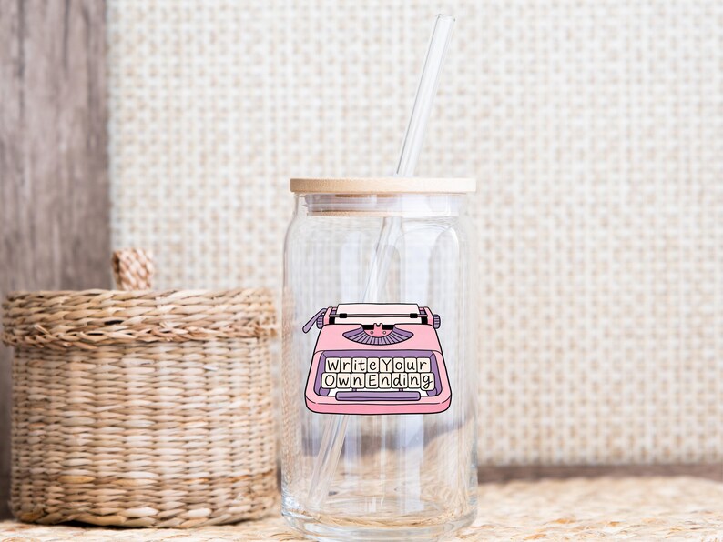 May include: A clear glass tumbler with a bamboo lid and a reusable straw. The tumbler features a pink typewriter graphic with the words "Write Your Own Ending". A woven basket and textured background are visible.