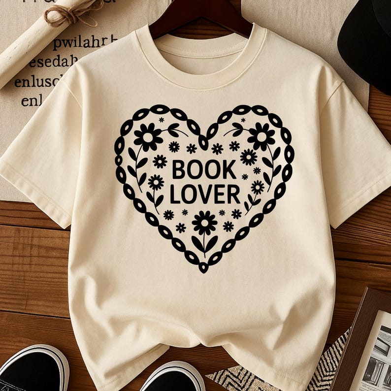 Puede incluir: Camiseta color crema con un dise&ntilde;o negro en forma de coraz&oacute;n con detalles florales y de hojas. Las palabras "BOOK LOVER" est&aacute;n centradas. La camiseta est&aacute; colgada en una percha.